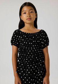 Schwarzes Oberteil mit weißen Punkten, das über einen gerafften Ausschnitt, kurze Ärmel und ein Peplum-Design mit gestuften Rüschen verfügt.