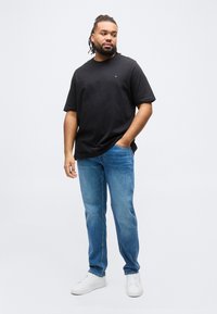 Άνδρας με πλεξούδες, που φοράει μαύρο μπλουζάκι, μπλε τζιν και λευκά αθλητικά παπούτσια, στέκεται με το ένα χέρι στην τσέπη σε απλό λευκό φόντο.