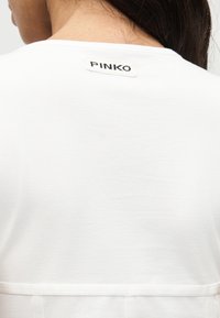 Πίσω όψη ενός ατόμου που φοράει λευκό πουκάμισο με μια μικρή ετικέτα «PINKO» κοντά στη λαιμόκοψη.