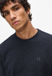 Dunkelblauer strukturierter Pullover mit Rundhalsausschnitt und dezentem Logoakzent in passender Farbe auf der Brust. Weiches Material.