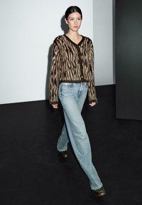 Cardigan a fantasia marrone e beige con bottoni, abbinato a jeans a gamba larga blu chiaro e scarpe scure. Modella che cammina su uno sfondo neutro.