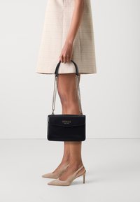 Borsa a mano in pelle nera con forma rettangolare, manico superiore e tracolla a catena. Presenta un logo dorato e una superficie liscia. Accompagnata da tacchi nudi.