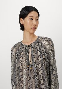 Python-print blouse met een losse pasvorm, geplooide halslijn en striksluiting. Kenmerkt zich door een zachte, doorschijnende stof in beige en zwarte tonen.