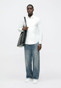 Balta pogājama krekls ar teksturētu virsmu, vaļīgas formas izbalējušas zilas džinsas un balti sporta apavi. Melna tote soma pār plecu.