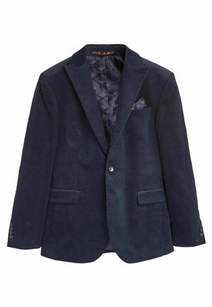 Mann trägt einen schwarzen Cord-Blazer über einem weißen T-Shirt mit schwarzer Hose und steht drinnen.