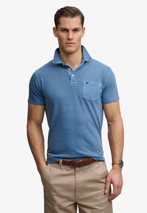Hombre con polo azul de manga corta con bolsillo en el pecho, pantalones beige, cinturón trenzado marrón y reloj de pulsera, de pie con las manos en los bolsillos.