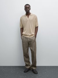 Béžový pletený polo svetr s krátkými rukávy, kombinovaný se světle hnědými lněnými kalhotami a černými tkanými sandály, na jednobarevném šedém pozadí.