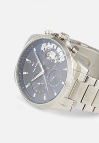 Silver metal watch with a black face, featuring three sub-dials and day/date display. Stainless steel band with interlocking links.