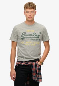 Camiseta gris de manga corta con texto impreso en verde y amarillo, "Vintage SuperDry", con una camisa de cuadros atada a la cintura.