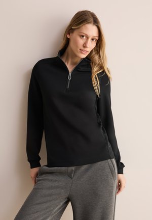 Schwarze Zip-Hoodie mit Stehhals, langen Ärmeln und glatter Textur, kombiniert mit grauen Sweatpants mit Seitentaschen.