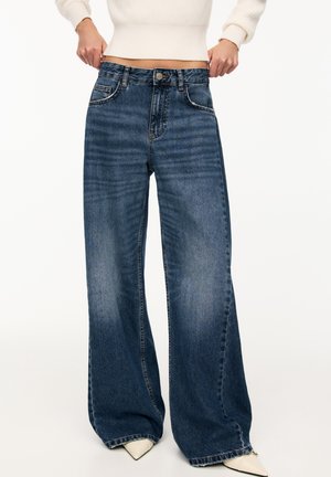 Personne remontant un jean large bleu foncé, associé à des chaussures blanches pointues et un pull court crème.