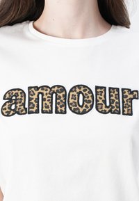 White cotton t-shirt featuring the word "amour" in bold, leopard print letters outlined in black. Short sleeves and crew neck design.