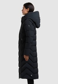 Kvinde stående i profil iført en lang sort quiltet vinterjakke med stor hætte og lynlåslomme i siden mod en ensfarvet baggrund.