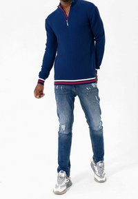 Pullover testo navy blu con colletto zip, caratterizzato da accenti a righe tricolori ai polsini e all'orlo, abbinato a jeans blu strappati e sneakers.