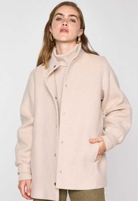 Cappotto in misto lana beige con una texture morbida, caratterizzato da un colletto alto, chiusure a pressione e tasche laterali. I polsini a coste completano il design.