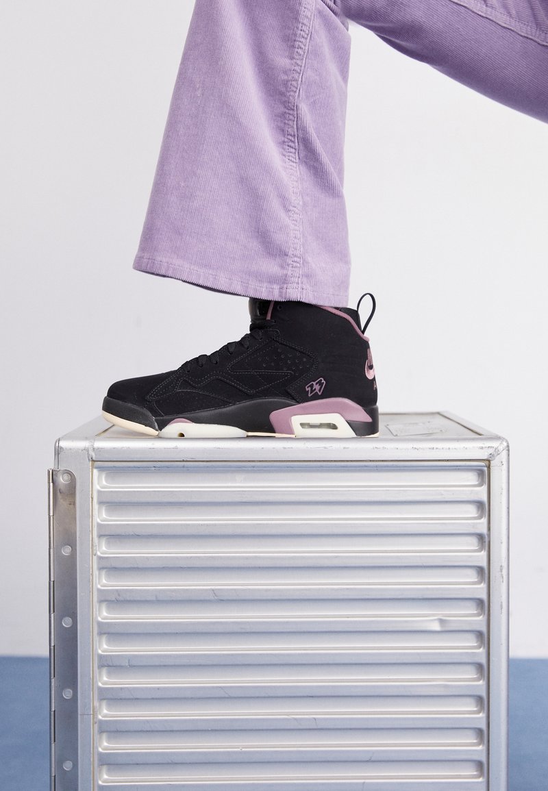 Personne portant des baskets noires et roses et un pantalon lavande, debout avec un pied sur une boîte de rangement métallique argentée.