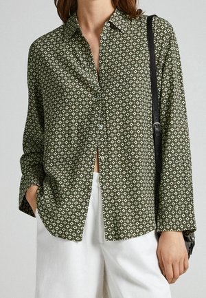 Camisa verde con botones y estampado de diseños geométricos blancos, mangas largas y un corte relajado. Combinada con pantalones blancos de pierna ancha.