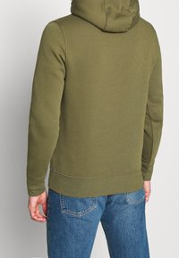 Felpa con cappuccio verde oliva, dal tessuto morbido, con polsini e orlo a coste. Indossata con jeans in denim azzurro chiaro, mostrando la vista posteriore.