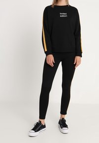 Sweat-shirt noir avec l'inscription « édition limitée » et des bandes latérales jaunes, associé à un legging noir et des baskets noires à semelles blanches.