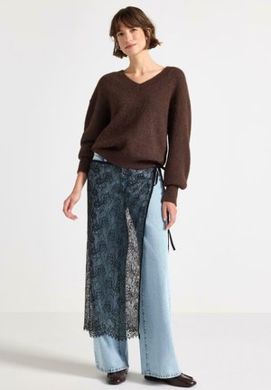 Donna che indossa un maglione marrone a scollo a V, jeans azzurri a gamba larga con sovrapposizione di gonna a portafoglio in pizzo nero trasparente e scarpe marrone scuro con punta quadrata.