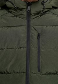 Chaqueta de invierno acolchada verde oliva con cremallera delantera negra, bolsillo en el pecho con cremallera y capucha con cordones en blanco y negro.