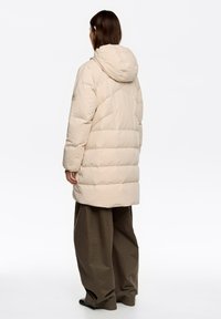 Chaqueta acolchada beige con capucha, diseño acolchado, mangas largas y ajuste holgado, combinada con pantalones verdes oliva sueltos.