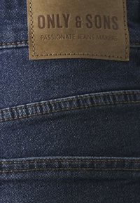 Mörkblå denim tyg med flera sömmar; läder etikett med "ONLY & SONS" och "PASSIONATE JEANS MAKERS" präglat.