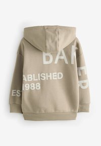 Bež hooded dukserica s velikim bijelim grafičkim tekstom: "BAKER", "OSNOVANO" i "1988" u različitim pozicijama. Mekani, teksturirani materijal.