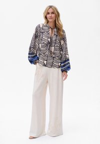 Blouse met abstract zwart en beige patroon, keyhole halslijn en blauwe ruches aan de mouwen, gecombineerd met beige wijde broek met bloemborduursel.