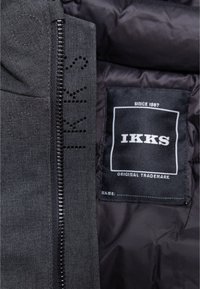 Donkergrijze jas interieur met een soepele textuur, voorzien van een zichtbare rits, geperforeerde details en een IKKS merklabel op de stof.