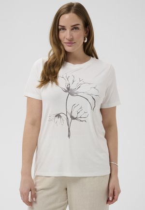 Femme aux longs cheveux bruns portant une chemise blanche à manches courtes avec un imprimé floral noir abstrait et un pantalon beige, debout devant un fond uni.