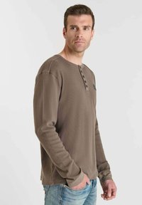 Hombre en una camiseta henley de manga larga marrón con tapeta de botones, con las manos en los bolsillos, mirando de lado contra un fondo simple.