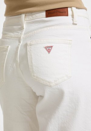 White denim jeans with a fitted design, featuring stitched pockets and a red triangular Guess logo on the back pocket. Brown leather waistband tag.