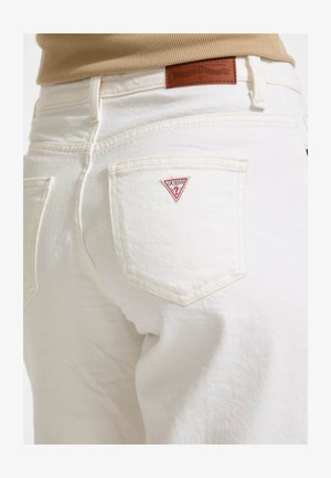 Bele kavbojke iz denima z oprijeto zasnovo, ki vključujejo šivane žepke in rdeči trikotni logotip Guess na zadnjem žepu. Oznaka iz rjavega usnja na pasu.