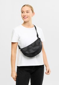 Zwarte met een patroon crossbodytas met een gebogen ontwerp, voorzien van een ritssluiting en een verstelbare schouderband. Draagt over een wit shirt.