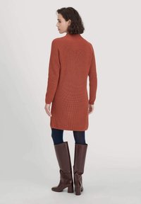 Frau, die mit dem Rücken steht und ein rostfarbenes Strickkleid, marineblaue Strumpfhosen und knielange, dunkle Lederstiefel trägt.