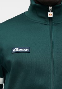 Πράσινο μπουφάν με φερμουάρ από υφασμάτινη υφή με ψηλό γιακά. Περιλαμβάνει λευκή μπαλώ patch με το λογότυπο 'ellesse' και φερμουάρ με επωνυμία.