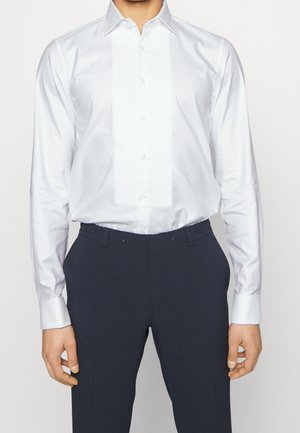 Homme portant une chemise blanche cintrée à manches longues et un pantalon formel foncé, debout avec les bras détendus le long du corps.