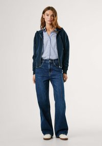 Vrouw in een marineblauwe zip-up hoodie, lichtblauw gestreept overhemd, wijde blauwe jeans en witte sneakers, staand tegen een neutrale achtergrond.