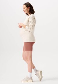 Graviditets sweatshirt i krämfärgad med långa ärmar, kombinerad med ribbade cykelshorts i rostfärg och ljusa sneakers med vita strumpor.