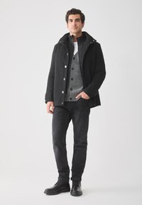 Hombre vestido con un abrigo negro, un cárdigan gris abotonado, pantalones negros y botas negras, de pie frente a un fondo claro y liso.