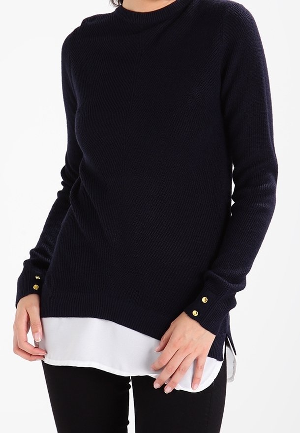 Marinblå ribbstickad tröja med långa ärmar, rund halsringning och guldknappdetaljer på ärmsluten, lager på en vit fåll.