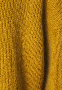 Gros plan sur un tissu tricoté moutarde texturé avec des fibres visibles et un pli vertical subtil.