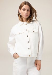 Witte denimjas met een spitse kraag, voorknopsluiting, twee borstzakken en bloemenborduurwerk op de linkerschouder.