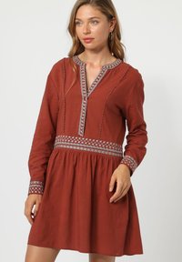 Vestido de color óxido con mangas largas, escote en V y bordados decorativos en blanco a lo largo del escote y los puños. Diseño de falda acampanada.