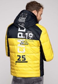 Zimowa kurtka w kolorze żółtym i czarnym, z kapturem, zamkiem błyskawicznym i różnymi tekstowymi grafikami. Teksturowany materiał i kontrastowe kolory.