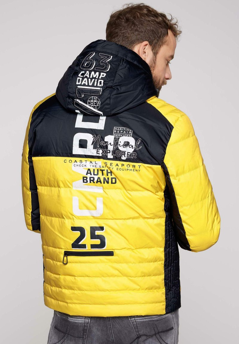 Zimowa kurtka w kolorze żółtym i czarnym, z kapturem, zamkiem błyskawicznym i różnymi tekstowymi grafikami. Teksturowany materiał i kontrastowe kolory.