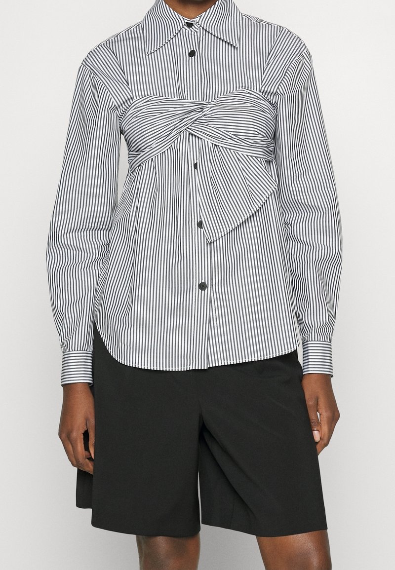 Shorts noirs associés à une chemise boutonnée à manches longues rayée verticalement en noir et blanc, avec un détail de tissu torsadé sur la poitrine.
