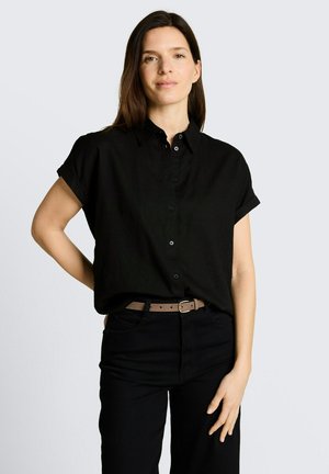 Woman wearing a short-sleeve black button-up shirt tucked into black pants with a beige belt, standing with one hand on hip.