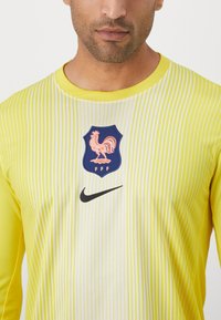 Dzeltena garām piedurknēm sporta krekls ar vertikālām baltām svītrām, ar emblēmu, kurā attēlots gailis un "FFF" logo; Nike swoosh melnā krāsā.